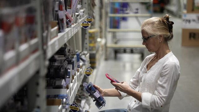 Mature woman using smart phone to take photo of door knobs for sale in a big box hardware store shopping for home renovation construction project.