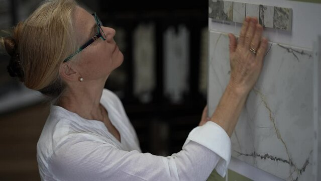 Closeup of mature woman touching ceramic tiles while shopping through samples for home redecorating renovation project, in a hardware store.