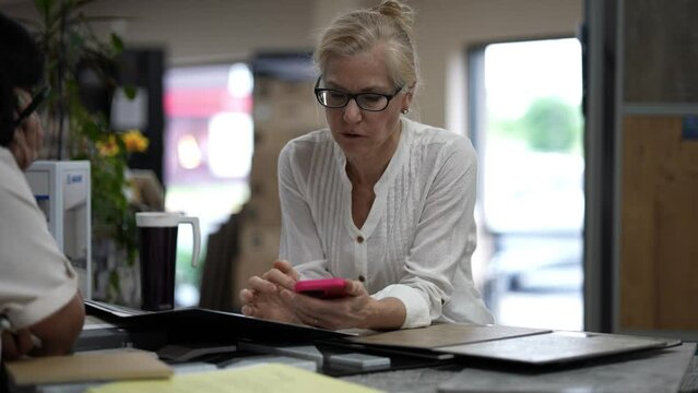 Mature woman holding smart phone talking to sales associate person regarding tiles to use for home renovation decorating project.