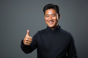 Fototapeta premium Portrait of a glad asian man in his 40s showing a thumb up while standing against minimalist or empty room background