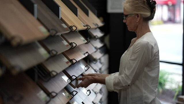 Happy mature woman looks at solid or engineered wood plank samples at a display in a floor or hardware store for a home remodeling renovation project.