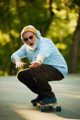 Elderly skateboarder cruises down park pathway, with headphones on, in stylish sunglasses and...