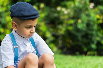 very sad little latino boy sitting on the ground in a public park looking down at the ground