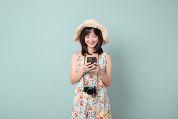 Travel and vacation concept. Happy Asian woman wearing casual dress and hat with camera while using mobile phone on vacation or travel theme isolated on pastel green background.