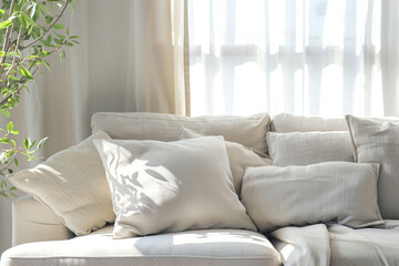 A serene living room with a plush beige sofa, surrounded by soft white pillows and curtains. The sunlight filters through the window creating gentle shadows on the fabric of the couch.