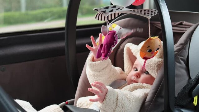 An infant lies in a car seat playing with a mobile, with a forest passing by in the background as the car moves. Close-up of the baby playing with the mobile