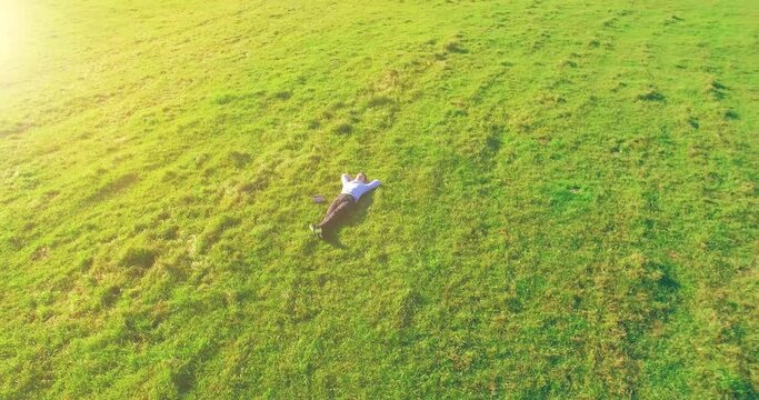 Aerial view point. Low orbital flight around man lied on green grass with notebook (pad) at yellow rural field. Casual weared. Sun rays. Radial movement.