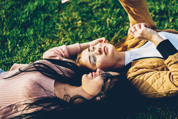 Two best friends bloggers in stylish sunglasses lying on green grass in park and making photos on...