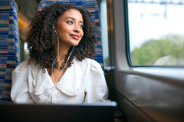 Businesswoman Commuting To Work On Train Working On Laptop Wearing Earphones Looking Out Of Window © Monkey Business