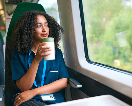 Female Doctor Or Nurse Commuting On Train Holding Reusable Coffee Cup Looking Out Of Window