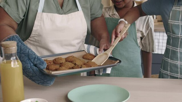 Midsection of African American juniors in aprons helping mom serve hot freshly baked oat cookies, carefully taking them off hot tray with wooden cooking spatula