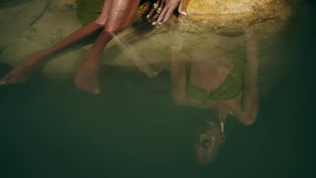 Reflection of androgynous black lgbtq fashion model lies on stones inside picturesque natural pool at night. Non-binary biethnic person poses in still water among rocks, touches water with hand