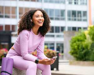 Woman Wearing Fitness Clothing Sitting Outdoors Streaming From Mobile Phone To Wireless Earbuds
