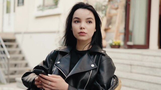 A young woman in a black leather jacket sits outdoors, holding and using her mobile phone. The image conveys a sense of casual and modern urban lifestyle. Young Woman Using Mobile Phone