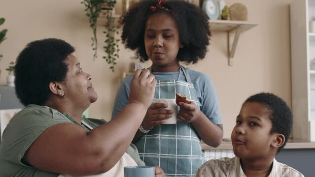 Medium of curly-haired African American siblings spending time with granny in kitchen, drinking tea and sharing freshly baked cookies with each other
