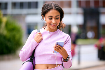 Woman Wearing Fitness Clothing Outdoors Listening To Music Or Podcast On Earphones From Mobile Phone