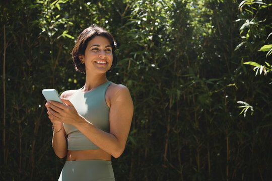 Happy Hispanic fit young woman using phone checking fitness applications standing in green nature park looking away advertising yoga retreat outdoor classes, mobile online sport apps. Copy space.