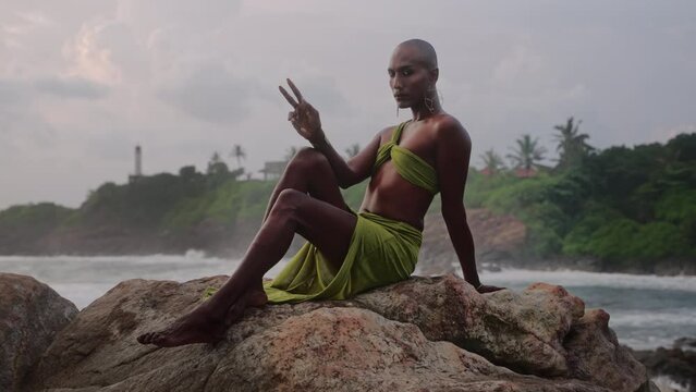 Androgynous person of color stands in boutique open dress on rock above ocean coastline on sunset. Trans sexual ethnic fashion model in skimpy outfit wears golden jewellery, posing like Oshun goddess.