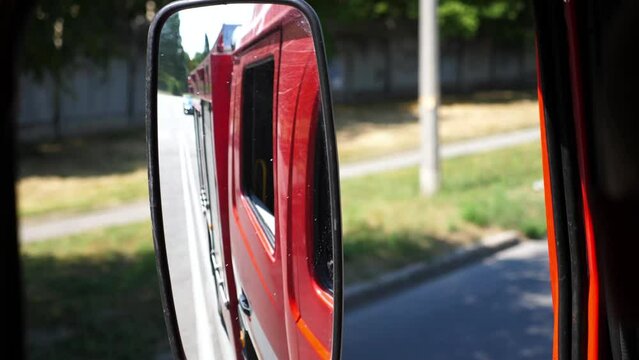 View to side mirror of fire engine riding to an emergency. Red big truck driving to place of a fire on sunny summer day. Concept of saving lives and heroic profession. Close up Slow motion