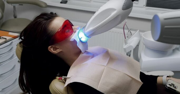 Teeth whitening procedure with ultraviolet light UV lamp in a modern clinic. Dental Clinic Teeth Whitening. Female patient in protective glasses at dentist chair. Whitening light bleaching woman teeth
