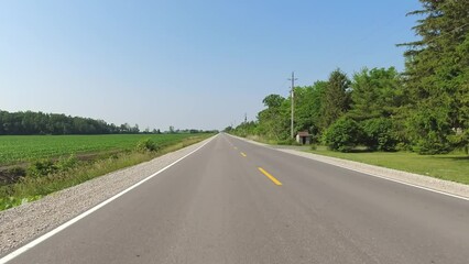 Driving in North America in the country side while adventures travel on vacation. Sun set at noon. Car back point of view shot at highway, farmer houses and farm fields Ontario province in Canada.