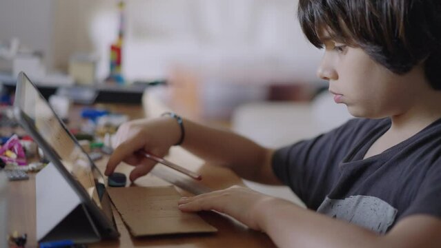 A young boy concentrates on a paper craft project while referring to instructions on a digital tablet at a table in a well-lit room.