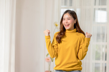 Happy woman in yellow sweater celebrating success indoors, showing joy and excitement with raised...