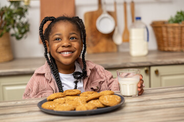 Joyful little girl enjoying delicious snack at home drinking milk with cookies