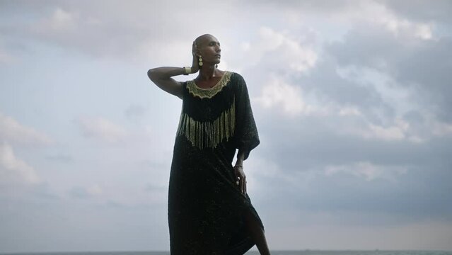 Androgynous person of color stands in boutique posh dress on rock above ocean coastline. Trans sexual ethnic fashion model in epic outfit wears golden jewellery, posing like Oshun goddess concept.