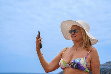 A mature caucasian woman wearing a hat and sunglasses taking a selfie with her mobile