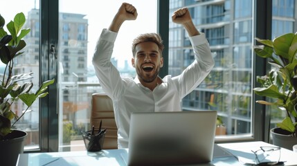 The handsome young businessman, sitting at his desk, using a laptop in the office, raises his hands and celebrates impressive fiscal results. The window in the background shows a cityscape view.