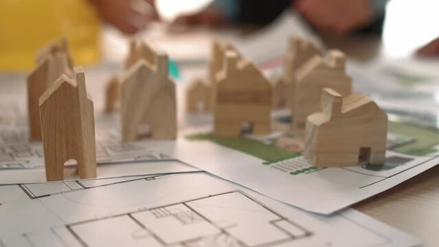 Architect and engineer discuss about building blueprint on table with architectural document, measurement tools scatter around at office, use wooden block for house positioning. Closeup. Delineation.