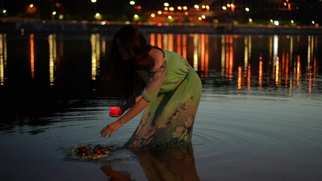 A mysterious girl in a green dress holds a burning candle in a candlestick in her hand while walking along a river knee-deep in the water. A girl poses on a night against the background of the lights