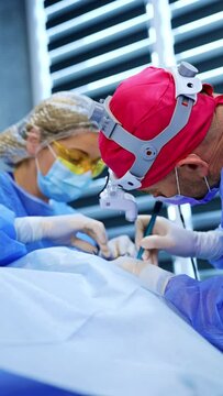 Caucasian male doctor and female nurse work at operation on thyroid. Chief doctor carefully uses surgical device at procedure. Vertical video.