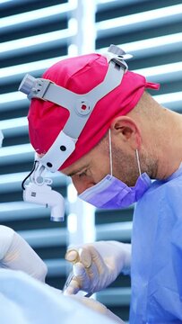 Male surgeon uses metal tools at operation on thyroid. Side view portrait of the doctor wearing pink cap and headlight. Vertical video.