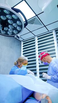 Surgeon wearing pink cap and headlights works in modern surgery room. Female nurse assists the doctor. Vertical video.