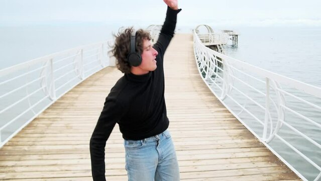 Cute young guy party-goer enjoying music in headphones, having fun dancing standing on the shore of the lake in happy solitude with his arms outstretched, celebrating acceptance to university.