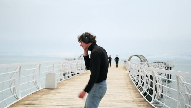 Fiery handsome guy dancing listening to music on headphones waving his arms standing on a beautiful lake bridge celebrating birthday against backdrop of people passing by in sunny weather.