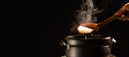 The cook is cooking rice porridge in a boiling clay pot with steam rising and using a wooden ladle...