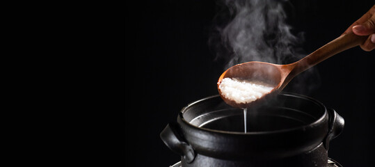 The cook is cooking rice porridge in a boiling clay pot with steam rising and using a wooden ladle...