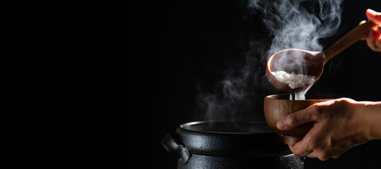The cook is cooking rice porridge in a boiling clay pot with steam rising and using a wooden ladle to scoop the rice porridge into a wooden bowl.