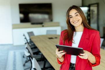 Smiling professional woman in red blazer holding tablet in modern conference room during daytime