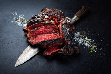 Barbecue dry aged chianina rib of beef steak served with crystal salt and thyme as close-up on a black design board with a large butcher knife