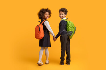 Two African American children, wearing backpacks, are holding hands against a bright yellow...