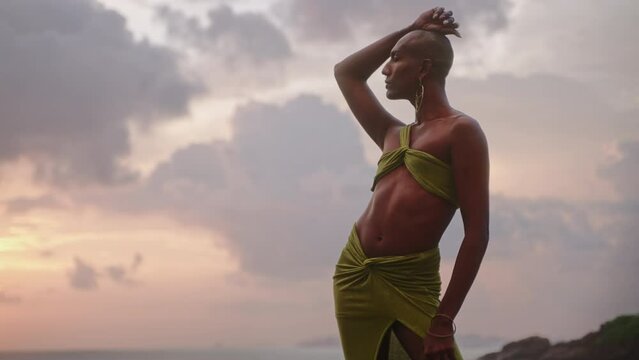 Androgynous person of color stands in boutique open dress on rock above ocean coastline on sunset. Trans sexual ethnic fashion model in skimpy outfit wears golden jewellery, posing like Oshun goddess.