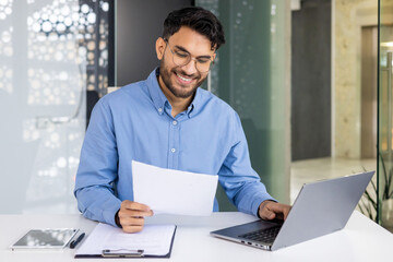 A smiling young Indian man is sitting in the office at a table with a laptop and working with documents, holding papers and typing data on a keyboard