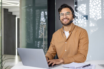 Portrait of a smiling young Muslim man wearing a headset, working in the office at a table on a...