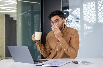 Ill Indian young man sitting in the office at the desk with a laptop, wiping his runny nose with a...