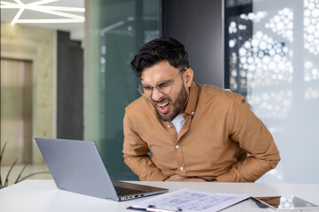 A young Indian man is sitting in the office, crouching in pain at his desk and holding his stomach