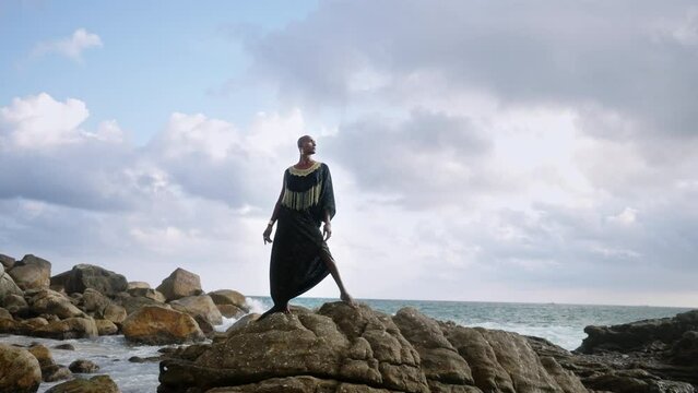 Gay bipoc man in luxury dress, boutique brass jewelry, makeup poses gracefully tropical rocky hill top on ocean beach. Homosexual ethnic elegant person standing on stone like Oshun orisha. Cloudy sky.